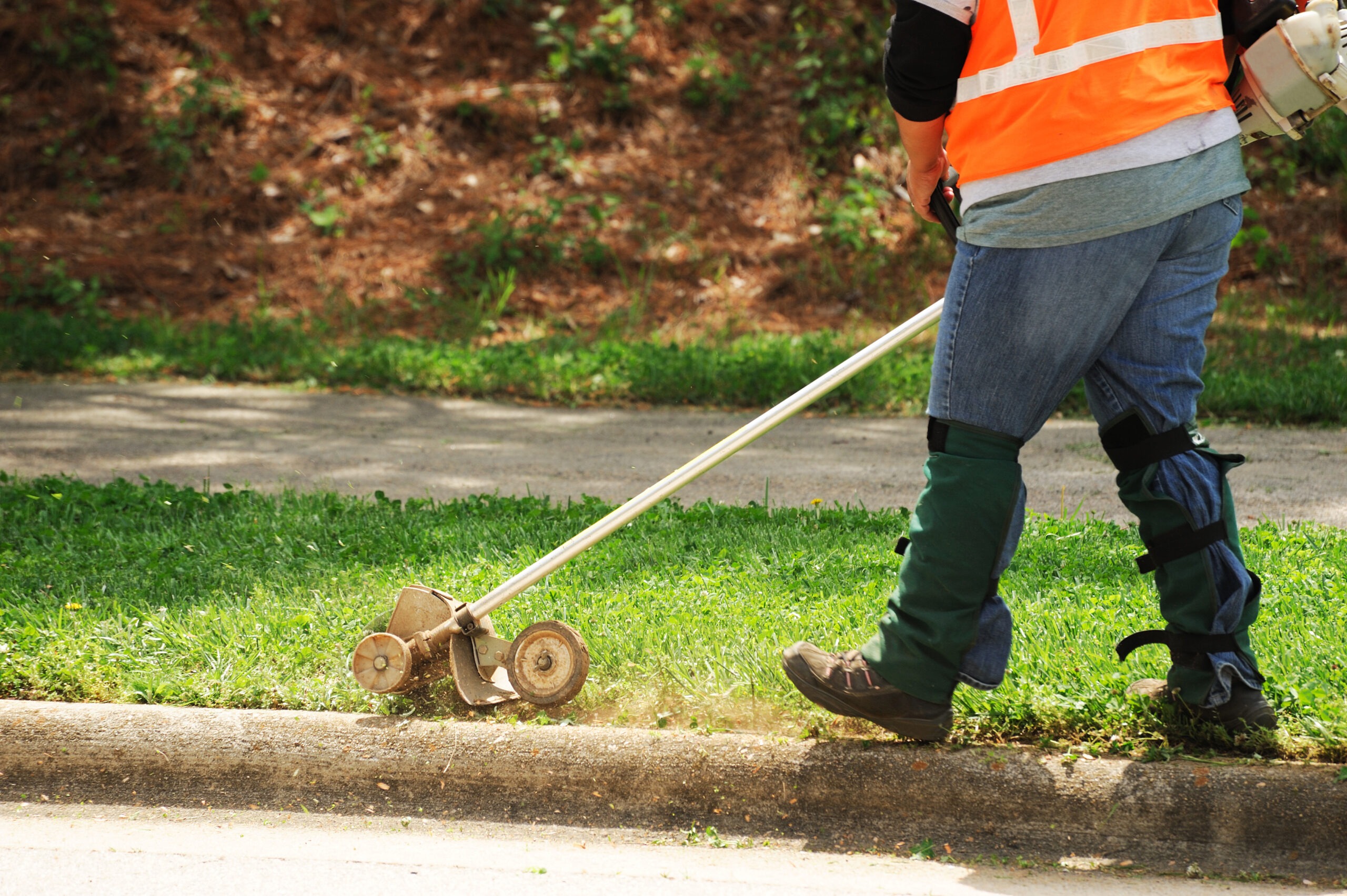 A worker in an orange safety vest uses a string trimmer to edge grass along a concrete curb